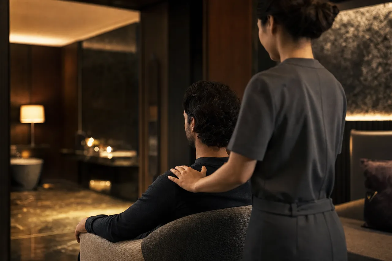 A relaxed male client seated upright in a warmly lit hotel suite, receiving a shoulder and neck massage from a therapist in a simple uniform